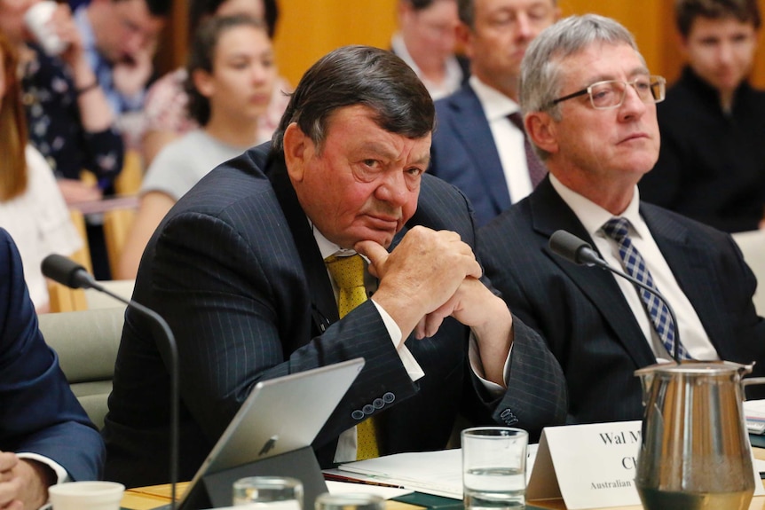 A man sitting at a desk with a crowd of people in a public gallery behind him