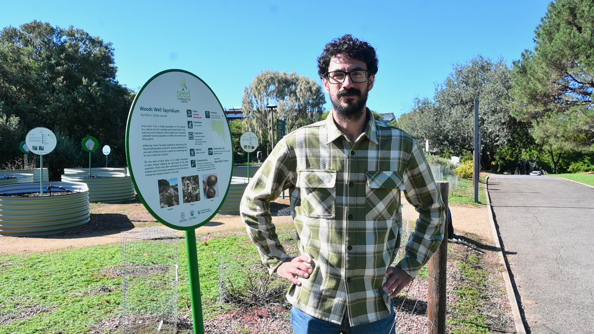 A man in a flannel shirt stands next to plants and signage