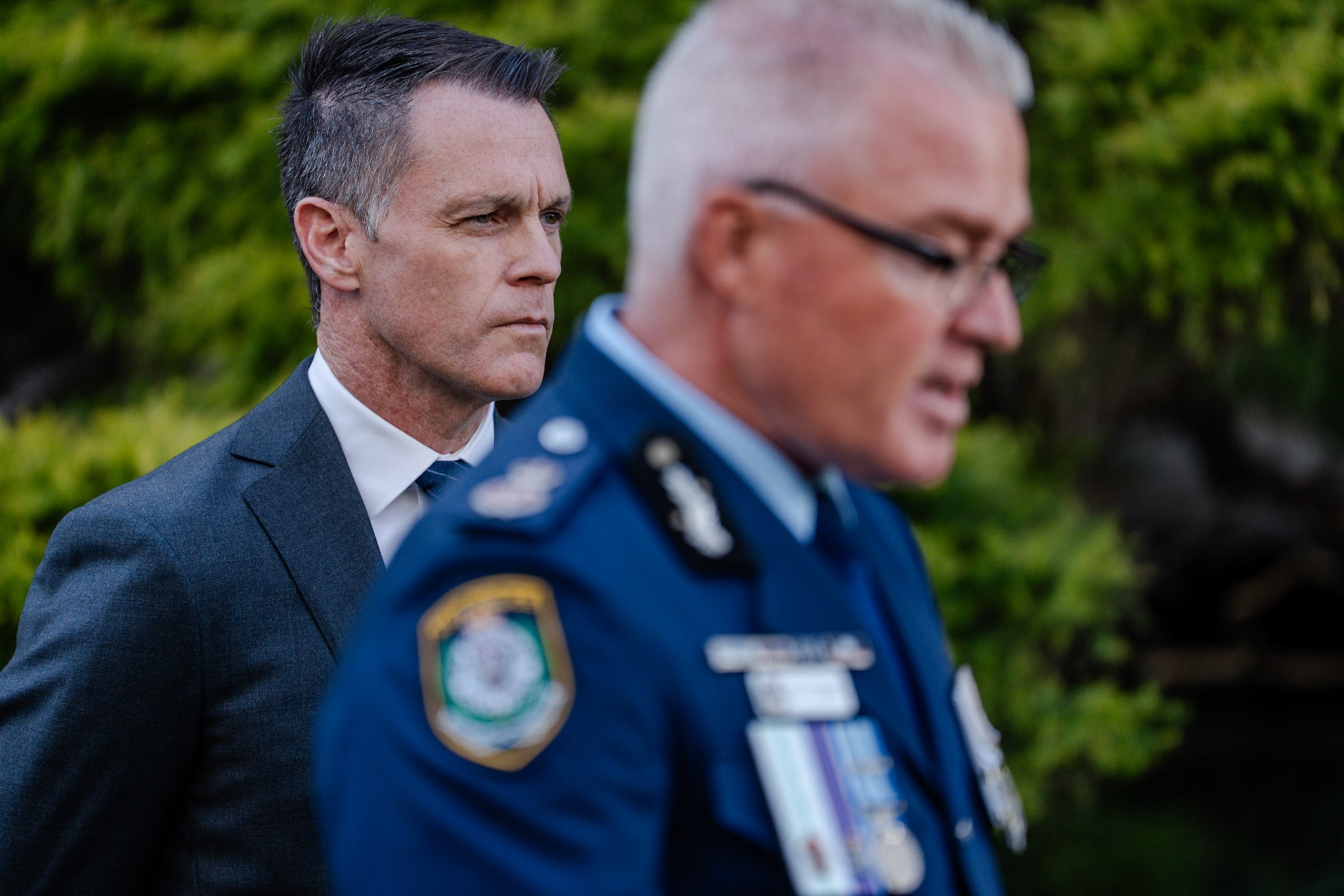 El primer ministro Chris Minns habla ante los micrófonos de los medios vestido con un traje gris, con Mal Lanyon y Yasmin Cately a su lado.