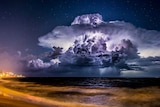 Lightning illuminates a cloud at night as a storm moves out to sea, with an illuminated beachfront also in shot.