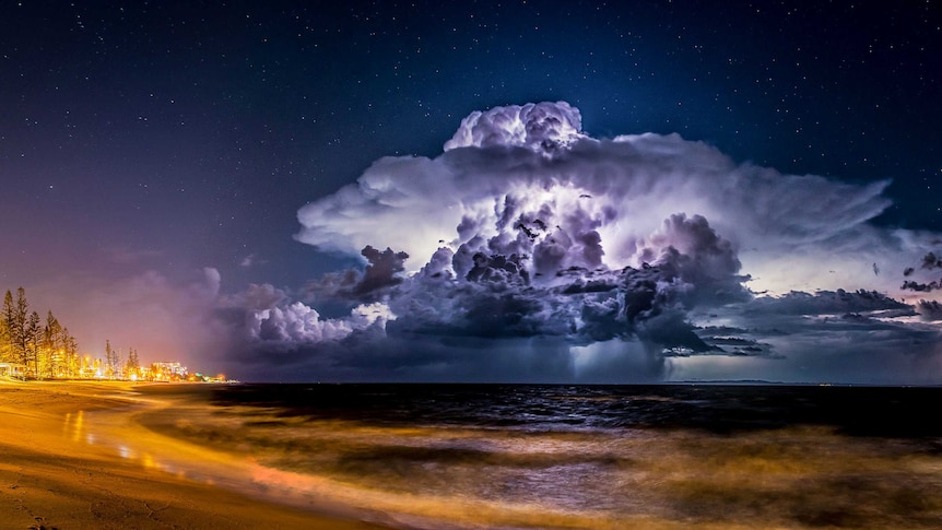 Lightning illuminates a cloud at night as a storm moves out to sea, with an illuminated beachfront also in shot.