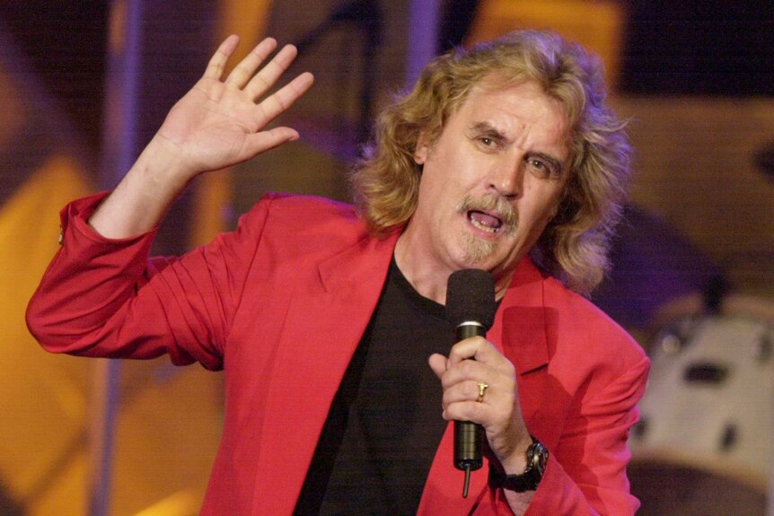 Billy Connolly holding a microphone and wearing a red jacket as he performs stand up.