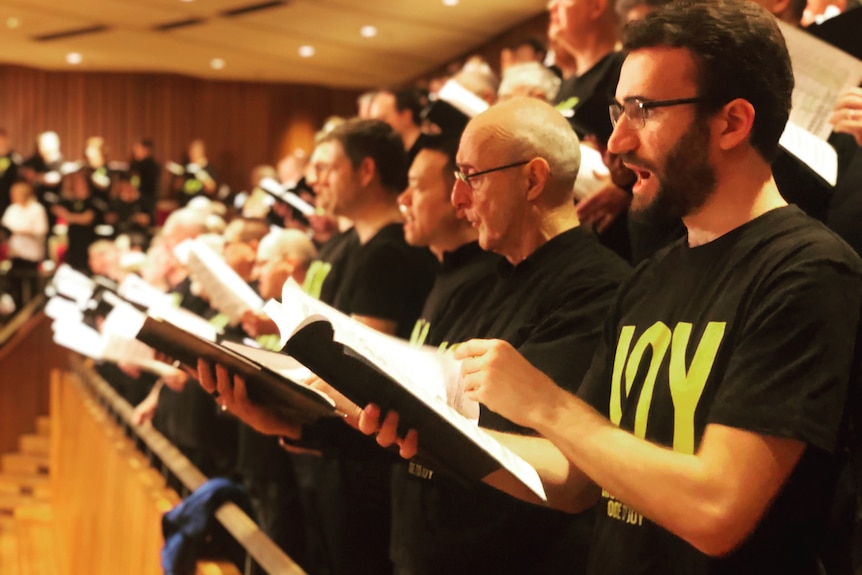 Close up shot of singers wearing 'joy' t-shirts, with mouths open mid-sing. Musicians are blurred in background.