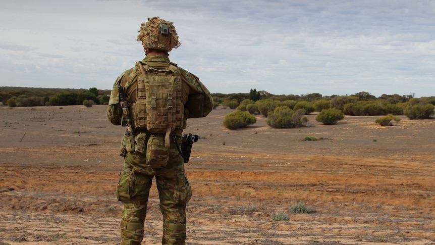 An Australian soldier in combat dress stands in an open field with his back to the camera.
