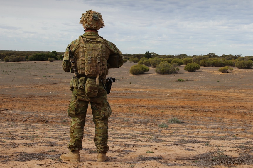 An Australian soldier in combat dress stands in an open field with his back to the camera.