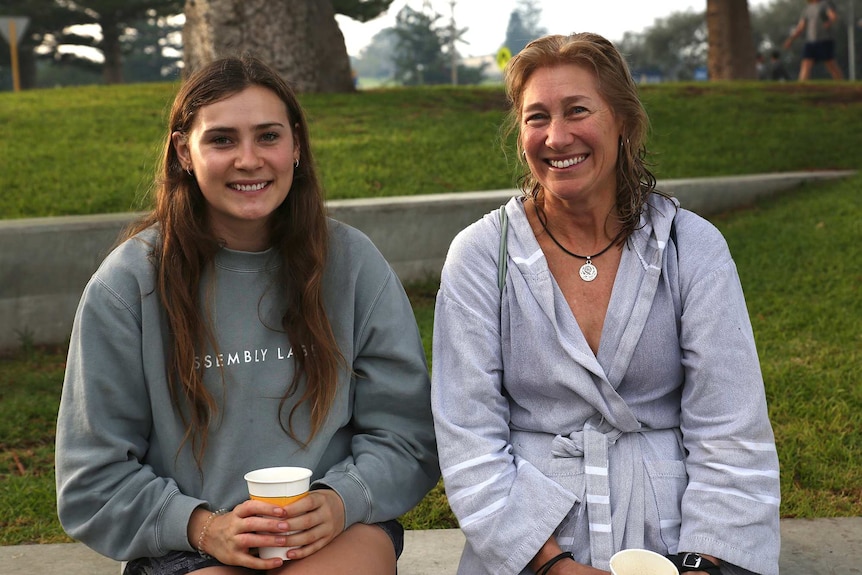 A daughter and her mother sit side by side smiling for a photo at Cottesloe Beach.