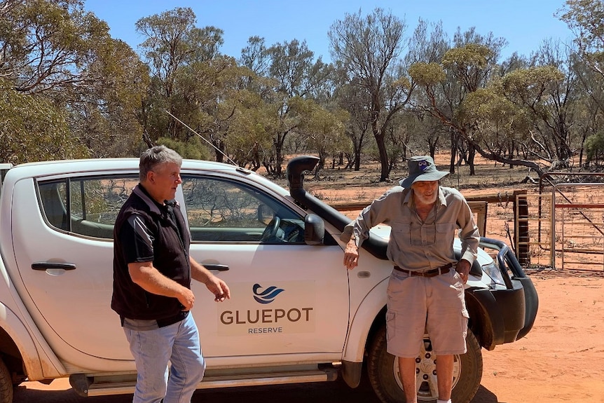 Two men stand in front of a white vehicle, looking away from the camera. There is orange sandy soil all around them.