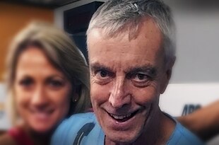 A lclose-up of a grey-haired smiling Ian, wears blue t-shirt, while a smiling loretta is blurred in the background.