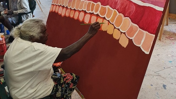 An elderly Indigenous woman sits down as she paints on a large red canvas