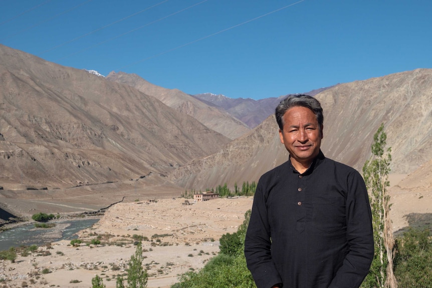 A man wearing a dark shirt and grey hair stands on some land near a mountain and blue sky.