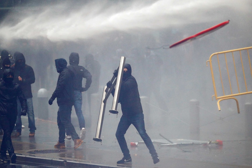 A man wearing a black hooded jacket throws a metal barricade as a water cannon sprays the crowd behind him