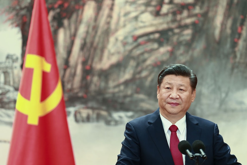 Xi Jingping stands behind a podium, a communist flag hangs behind him