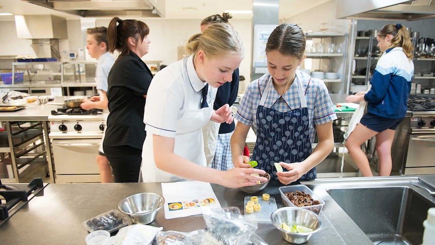 Two year 10 girls in school uniforms put food into bowls in a school kitchen.