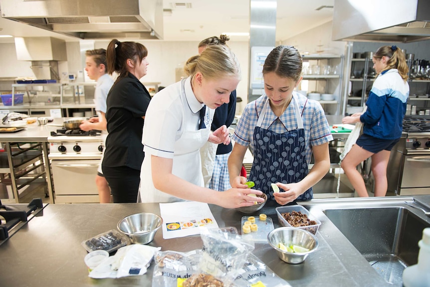 Two year 10 girls in school uniforms put food into bowls in a school kitchen.