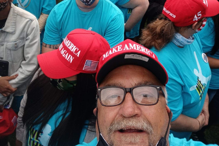 Randall Thom, in a crowd and wearing a MAGA hat, takes a selfie photo.