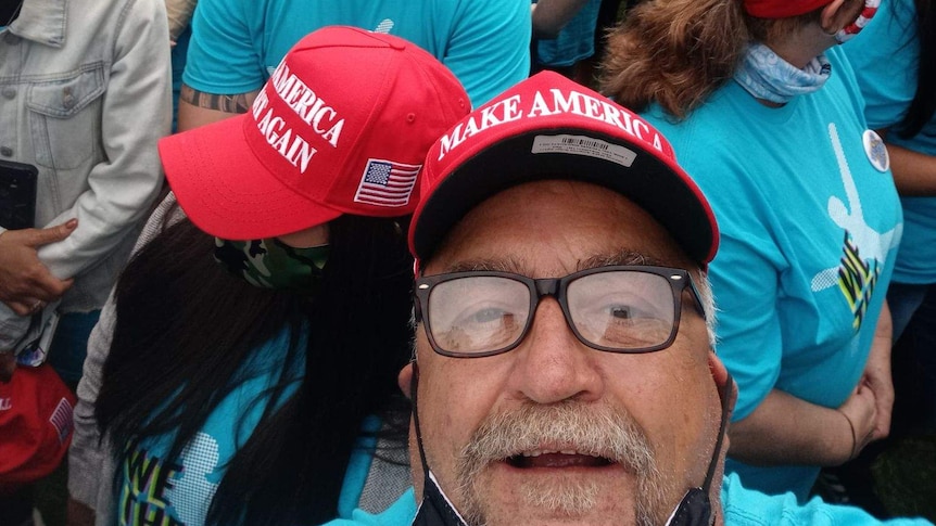 Randall Thom, in a crowd and wearing a MAGA hat, takes a selfie photo.