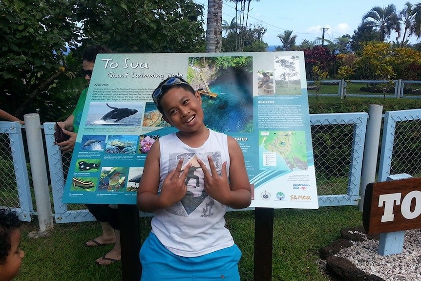 A young boy wearing a grass headband makes a sign with his hands as he is photographed in front of a tourist sign