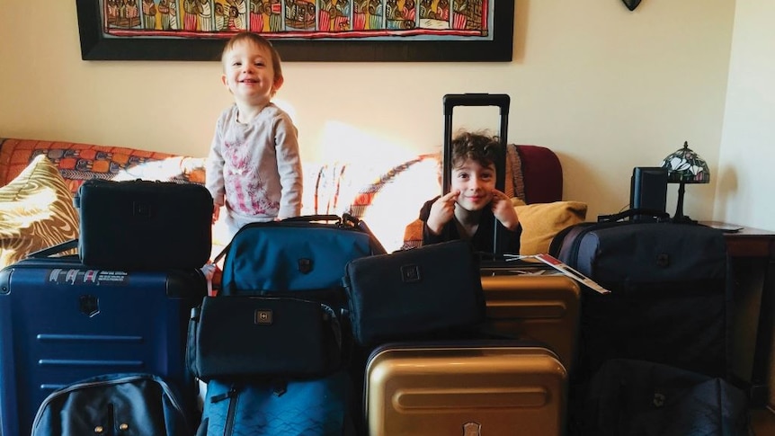 In a room filled with bags and suitcases, two small kids hide between them and smile mischievously.
