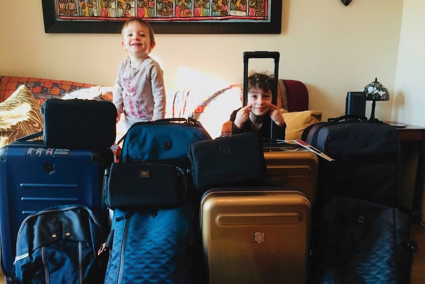 In a room filled with bags and suitcases, two small kids hide between them and smile mischievously.