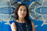 A brunette woman in her early 60s stands in front of a blue graphic print with white birds