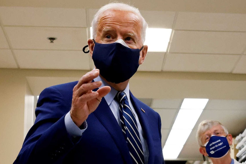 President Joe Biden in blue suit and tie gestures while health staff in background listen
