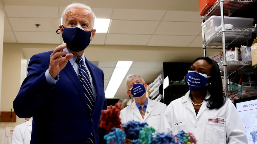 President Joe Biden in blue suit and tie gestures while health staff in background listen