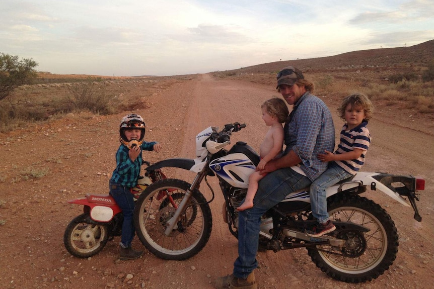 A man on a motorbike with two small boys, one in a nappy, mounted either side of him and another child on a smaller bike.