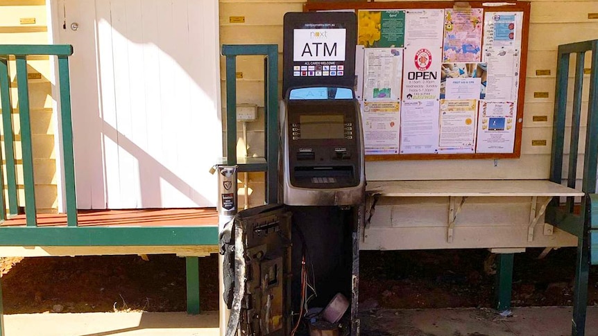 A blackened, destroyed ATM stands outside a yellow weatherboard building with a community noticeboard with colourful flyers.