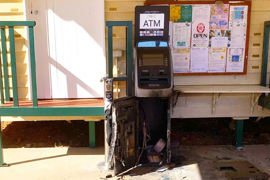 A blackened, destroyed ATM stands outside a yellow weatherboard building with a community noticeboard with colourful flyers.