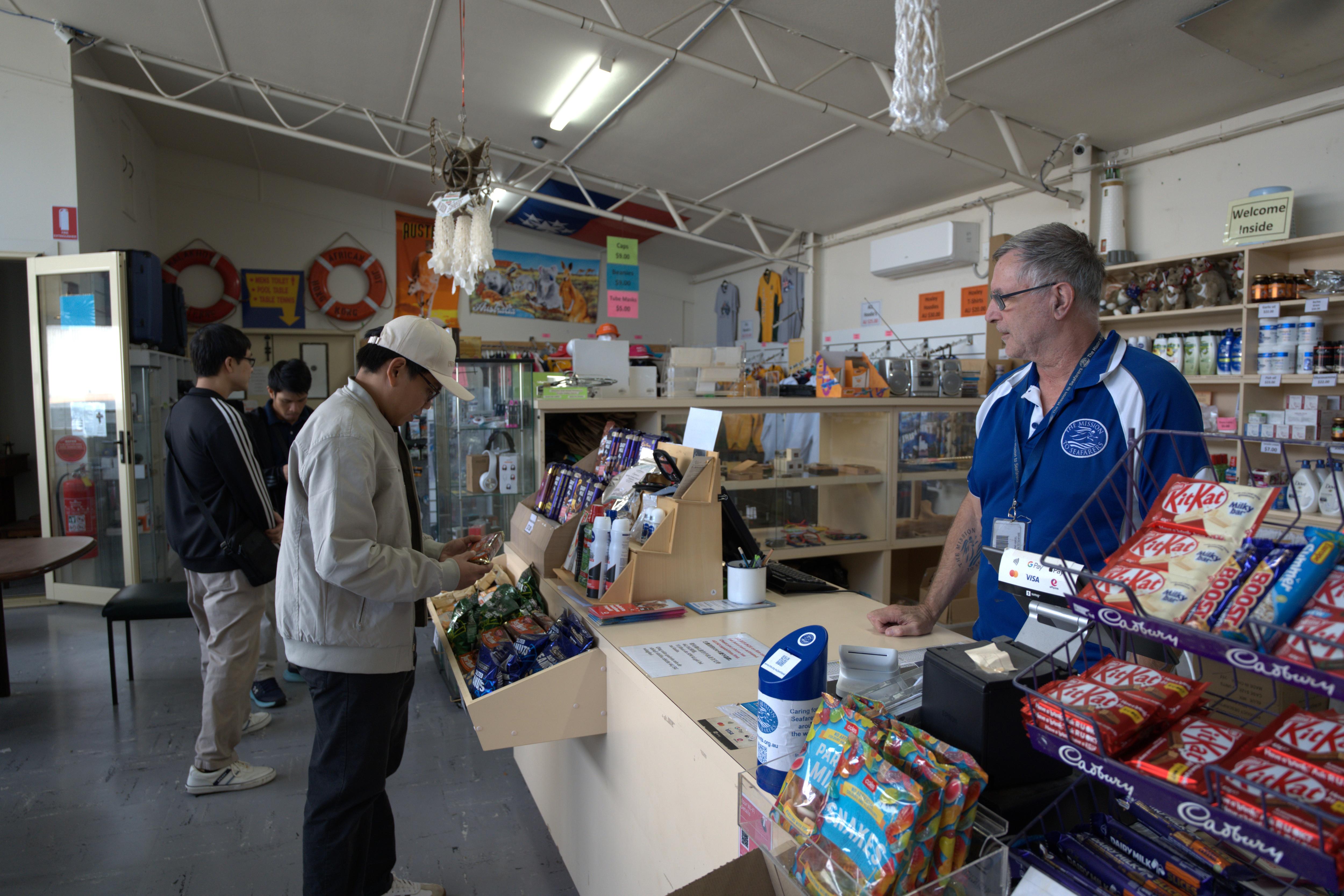A man is selecting a chocolate at the register of a shop, there are two other men browsing the shop behind him.