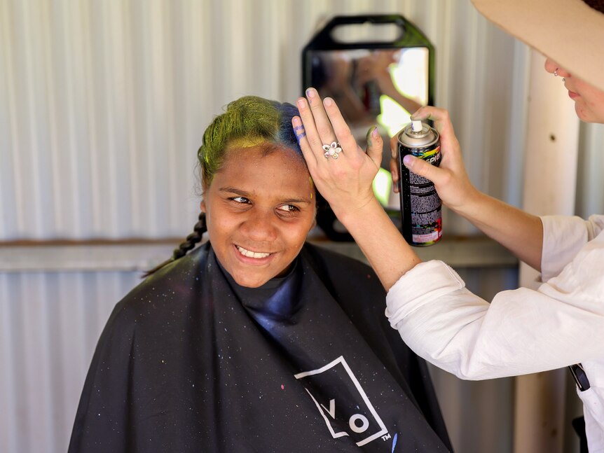 Young Aboriginal girl smiles as she has her hair sprayed yellow and blue by a white woman inside a shed