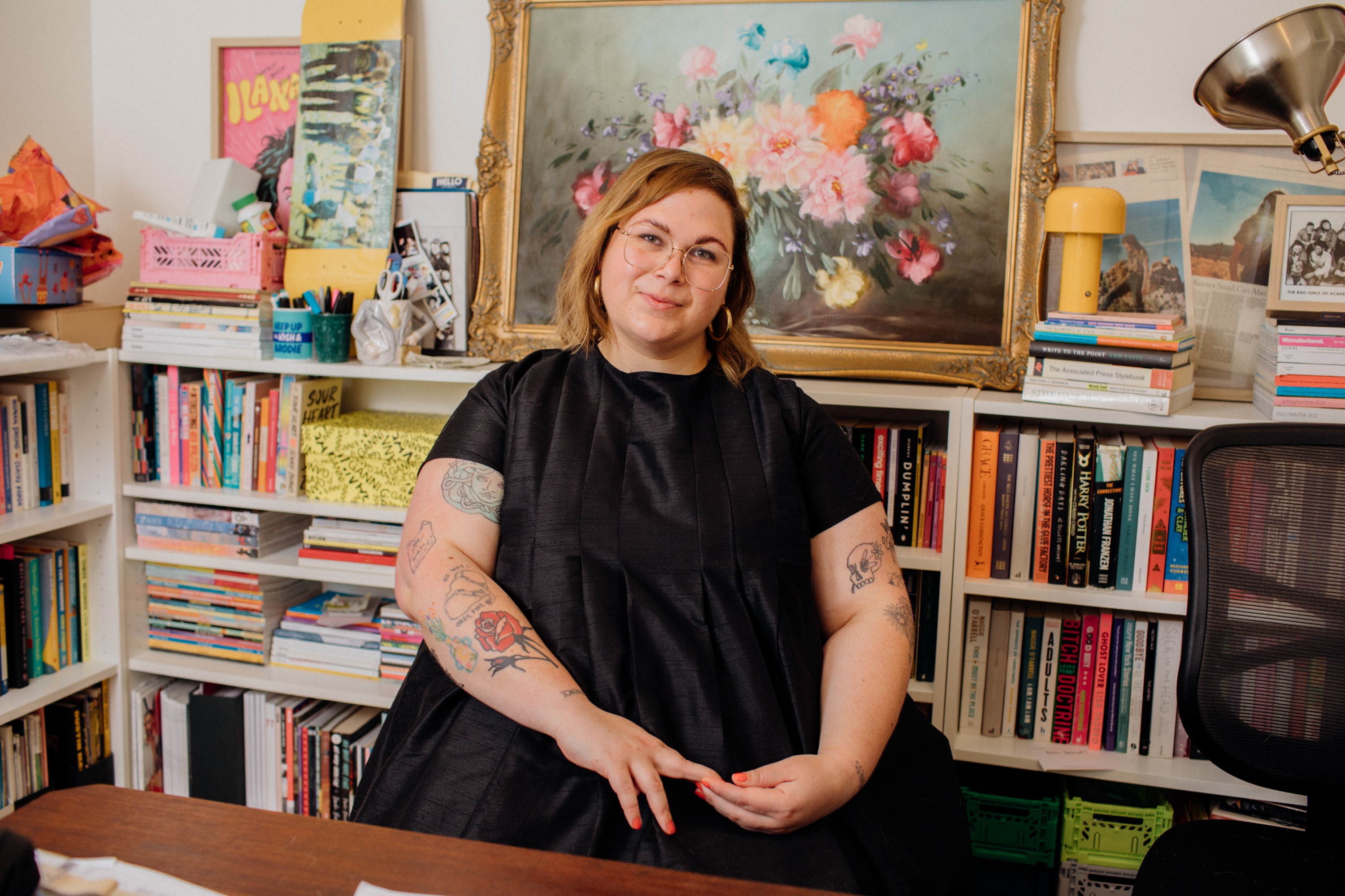 A woman in a Black dress and wearing glasses sits in front of a stacked bookshelf, topped with colourful artworks.