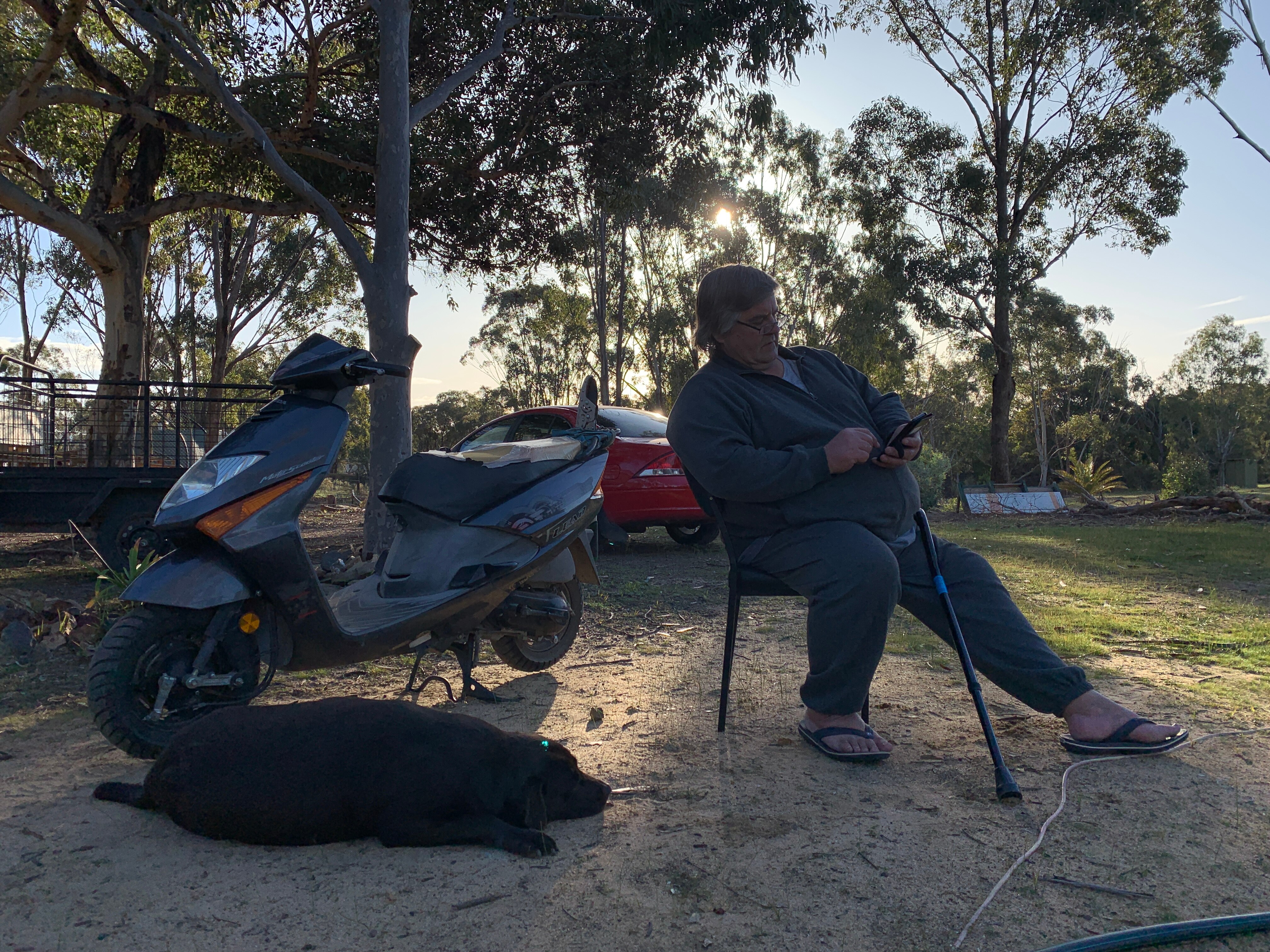 Man sitting outside next to a motorbike holding a walking stick.