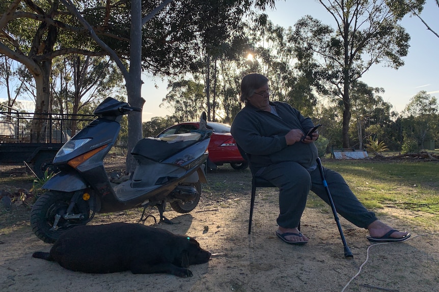 Man sitting outside next to a motorbike holding a walking stick.