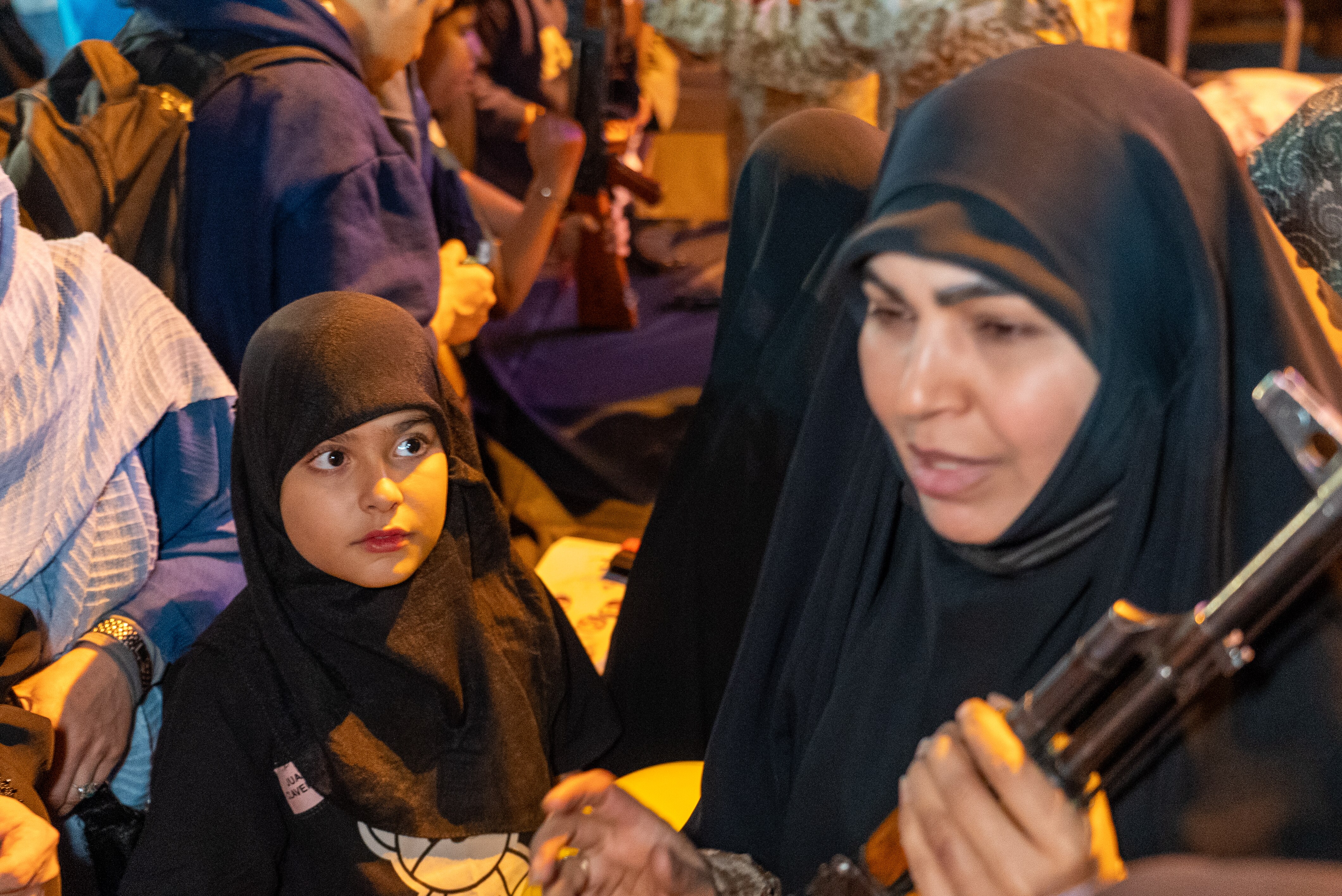 A young girl in traditional Islamic attire looking at a woman holding a rifle.