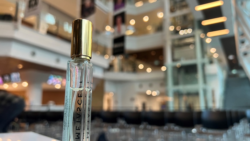 A small bottle of perfume with a gold lid with the lobby of a building in the background. 