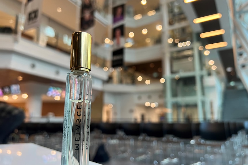 A small bottle of perfume with a gold lid with the lobby of a building in the background. 