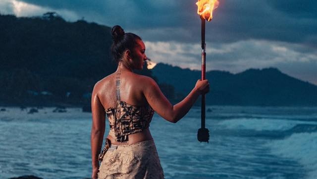 Moemoana Schwenke stands on a rock in front of the ocean holding a large machete on fire.