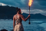 Moemoana Schwenke stands on a rock in front of the ocean holding a large machete on fire.