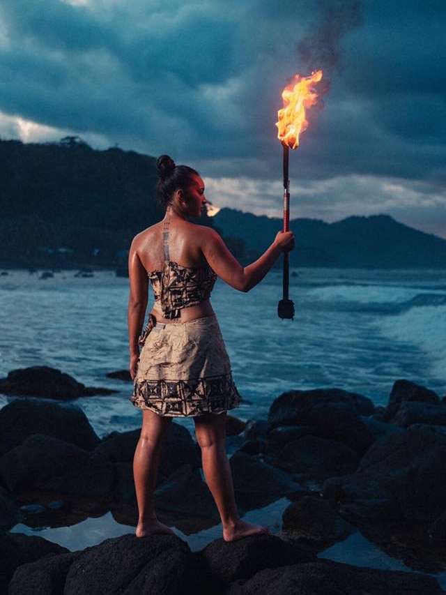 Moemoana Schwenke stands on a rock in front of the ocean holding a large machete on fire.