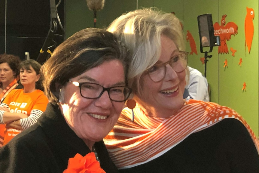 Two women stand together, dressed in black with orange accessories, smiling.