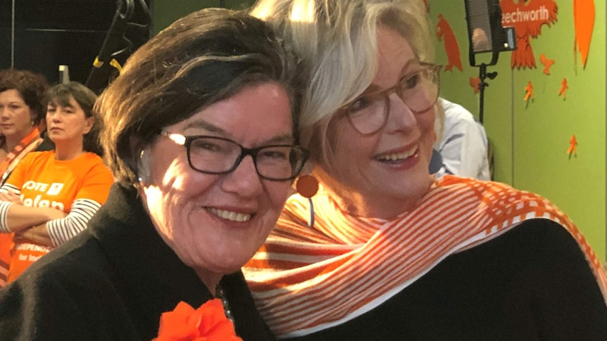 Two women stand together, dressed in black with orange accessories, smiling.