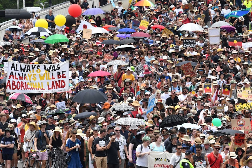 Thousands of people are seen crossing Victoria Bridge with placards, flags and umbrellas to protest Australia Day.