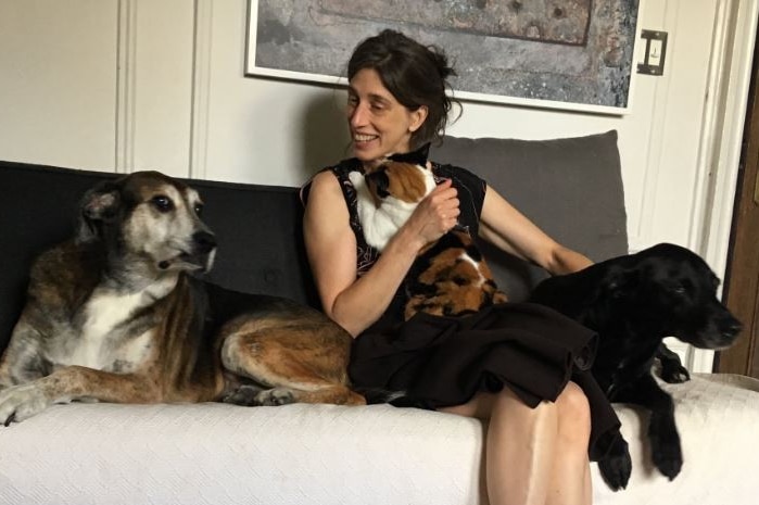 A smiling woman in black dress sits on couch with cat and dog on her lap and large dog at her side on the couch.
