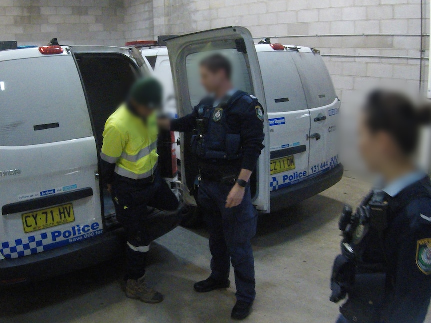A man in handcuffs is escorted by two police officers out of a police van.