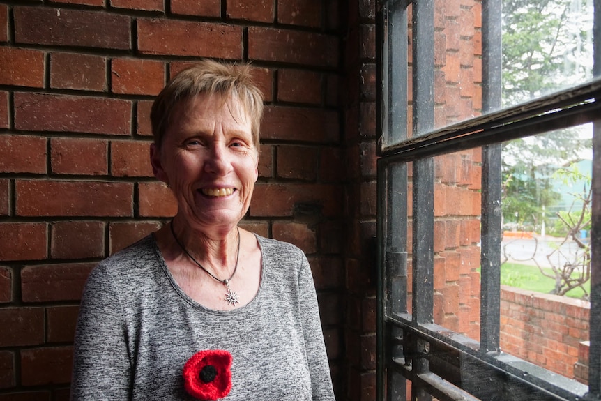 A woman in a grey shirt with a knitted poppy on it stands inside a brick building next to a window.