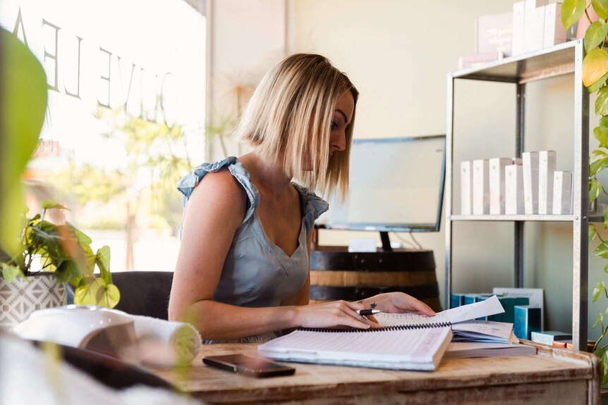 A woman with blonde haair and a blue top sits at a desk doing paperwork.