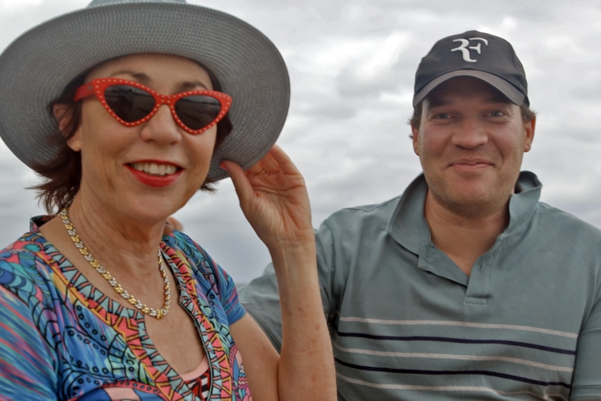 A woman with short brown hair holding a hat on her head on a windy day and red sunglasses sits next to her smiling adult son 
