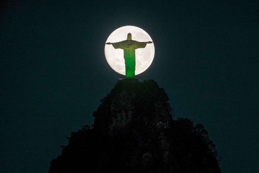 The moon descends behind the statue of Christ the Redeemer in Rio, lit by the colours of Brazil.