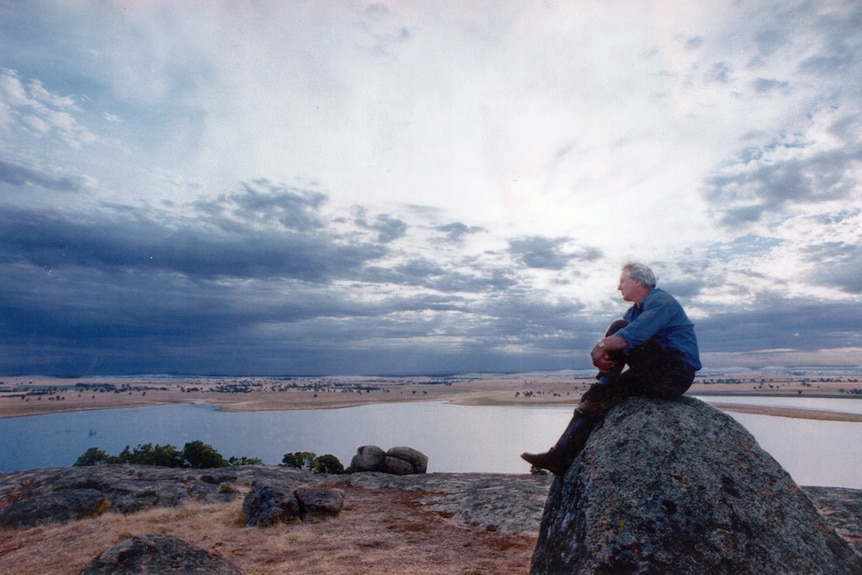 Rai sits on a boulder looking across a lake and cloudy sky. There are smaller rocks and grass in the foreground.